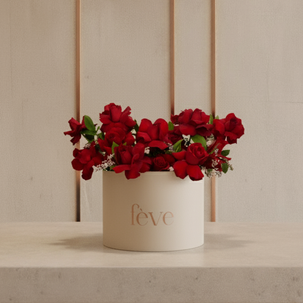 A cream-colored cylindrical box with the word “fève” in rose-gold lettering holds an arrangement of deep red roses accented with small white filler flowers and green leaves.