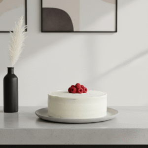 A smooth, white-frosted cake sits on a gray plate atop a light countertop. A small cluster of fresh raspberries is arranged at the center of the cake.