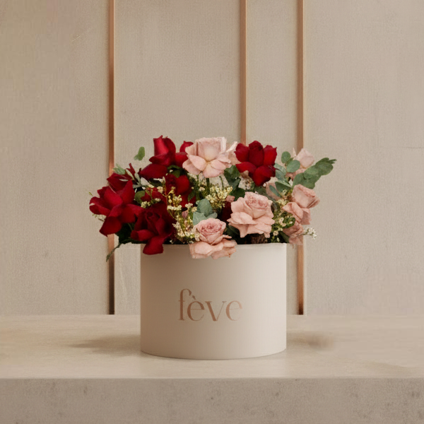 A round, cream-colored box with the word “fève” in rose-gold lettering holds an arrangement of roses in rich red and soft pink tones, accented with greenery and small white filler flowers.