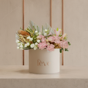 A cream-colored cylindrical box with the word “fève” in rose-gold lettering contains an arrangement of soft pink flowers, white blooms, dried grasses, and decorative fan-shaped leaves.