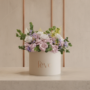 A cream-colored cylindrical box with the word “fève” in rose-gold lettering holds an arrangement of soft pastel flowers, including pale pink roses, white blooms, and small purple blossoms, accented with lush greenery.