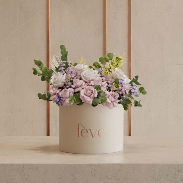 A cream-colored cylindrical box with the word “fève” in rose-gold lettering holds an arrangement of soft pastel flowers, including pale pink roses, white blooms, and small purple blossoms, accented with lush greenery.