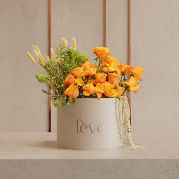 A cream-colored cylindrical box with the word “fève” in rose-gold lettering holds a vibrant arrangement of bright orange roses, green blooms, soft white accents, and cascading ivory amaranthus.