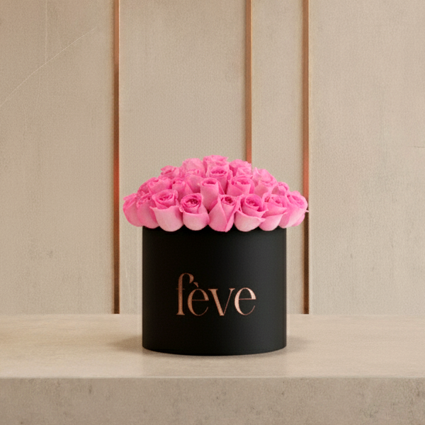 A black cylindrical box with the word “fève” in rose-gold lettering is filled with a tightly arranged dome of bright pink roses.
