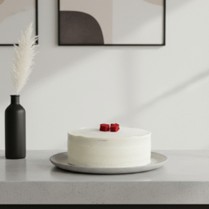 A smooth, white-frosted cake sits on a gray plate atop a light countertop. Two small red cake cubes are placed at the center of the cake as decoration.
