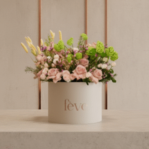 A cream-colored cylindrical box with the word “fève” in rose-gold lettering holds an abundant floral arrangement featuring soft pink roses, delicate white blooms, bright green hydrangea-like clusters, and accents of purple flowers and airy foliage.