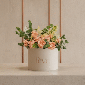 A cream-colored cylindrical box with the word “fève” in rose-gold lettering holds an arrangement of soft peach roses, airy yellow sprigs, and lush green foliage.