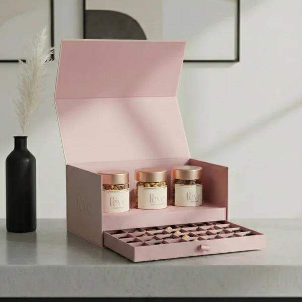Luxury pastel pink gift box by Feve featuring three jars of gourmet treats and a pull-out drawer of assorted chocolate truffles in a minimalist setting.