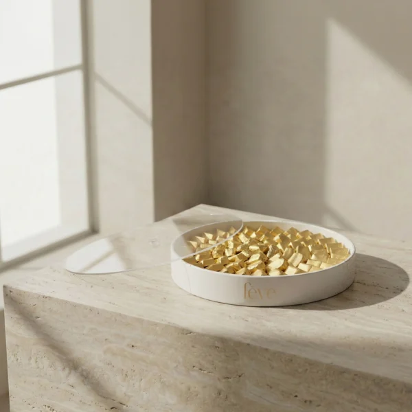 L’Orbe luxury chocolate tray by fève. Minimalist stone-textured circular vessel filled with gold-foil wrapped artisanal chocolates on a travertine plinth. High-end confectionery gift set with clear acrylic lid.