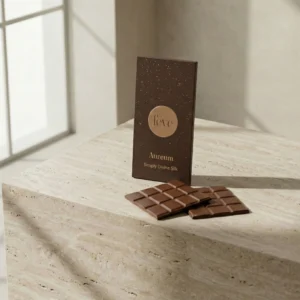 A luxury fève Aureum milk chocolate bar in dark brown and gold packaging, standing on a light-colored travertine stone surface next to two broken squares of chocolate.