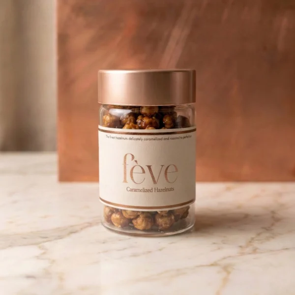 Gourmet fève caramelized hazelnuts in a luxury short glass jar with a rose gold metallic lid, presented on a marble surface.