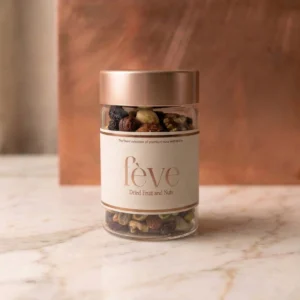 A minimalist glass jar of fève Dried Fruit and Nuts featuring a premium cream label with rose-gold lettering. The jar is filled with a gourmet mix of walnuts, almonds, and dark raisins, topped with a metallic copper lid. It is set against a warm, textured copper backdrop on a white marble surface, evoking a sense of modern luxury.