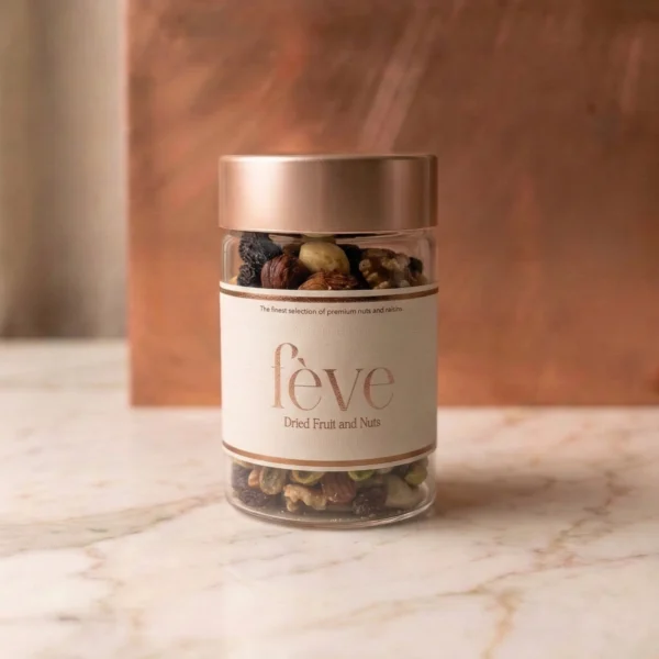 A minimalist glass jar of fève Dried Fruit and Nuts featuring a premium cream label with rose-gold lettering. The jar is filled with a gourmet mix of walnuts, almonds, and dark raisins, topped with a metallic copper lid. It is set against a warm, textured copper backdrop on a white marble surface, evoking a sense of modern luxury.