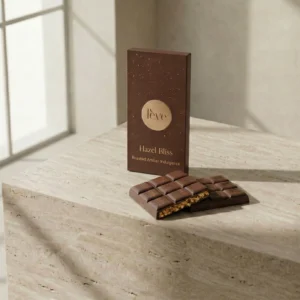 A luxury dark brown chocolate box labeled 'fève Hazel Bliss' standing on a textured travertine stone surface. In front of the packaging, broken pieces of milk chocolate reveal a dense layer of crushed roasted hazelnuts inside, set against a soft, sunlit minimalist background.