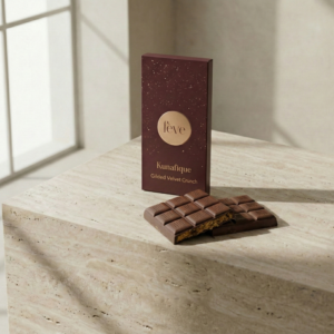 A luxury Kunafique milk chocolate bar by Fève, featuring a broken section revealing a crispy, golden toasted kataifi kunafa filling on a minimalist travertine stone surface.