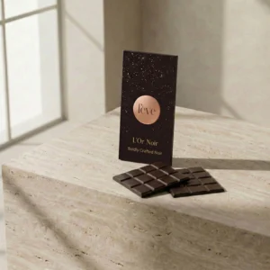 A sleek, dark chocolate bar titled "L'Or Noir" by Fève, standing on a textured stone surface next to two broken squares of dark chocolate in a minimalist, sunlit room.