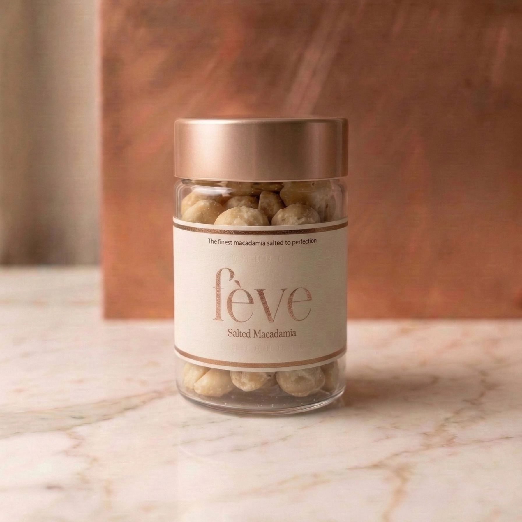 The jar features a minimalist cream-colored label with elegant rose-gold metallic typography. It is topped with a matching brushed rose-gold lid. The macadamias are visible through the glass, showing a light, salted dusting. The jar sits on a polished white marble surface against a warm, textured copper or rose-gold background, creating a luxurious and sophisticated aesthetic.