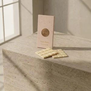 Artisanal Fève Satin Blanc white chocolate bar in speckled pink packaging and ivory chocolate squares on a textured travertine stone pedestal.