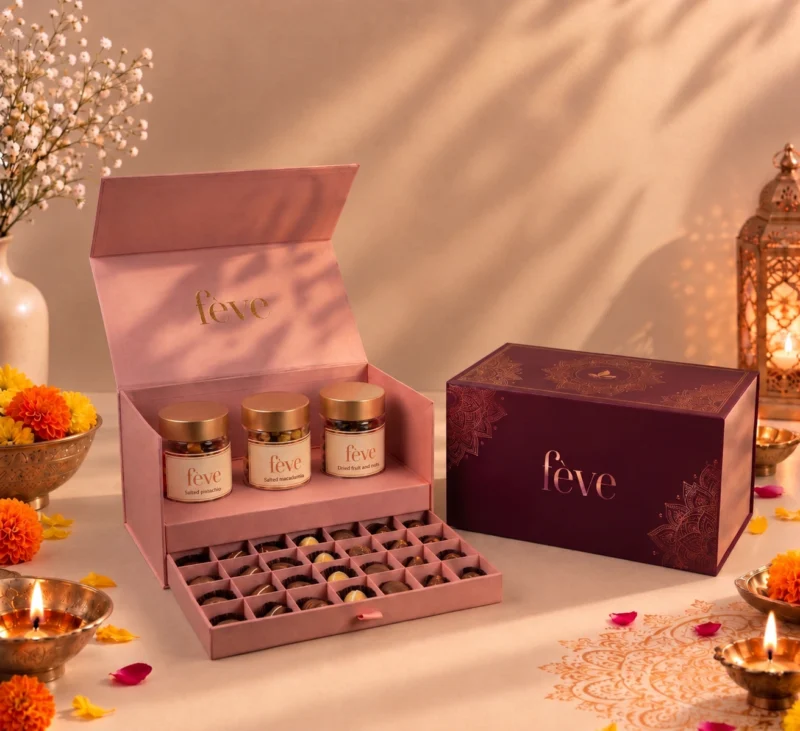Luxury fève Diwali gift set featuring an open pink dual-tier chocolate box with three nut jars and assorted chocolates alongside a maroon gold-accented gift box, surrounded by lit diyas and marigolds.
