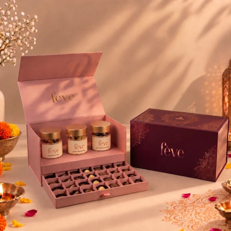 Luxury fève Diwali gift set featuring an open pink dual-tier chocolate box with three nut jars and assorted chocolates alongside a maroon gold-accented gift box, surrounded by lit diyas and marigolds.
